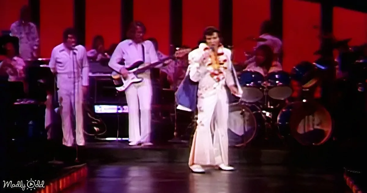 A performer in a white suit with a colorful sash sings on stage with a band.
