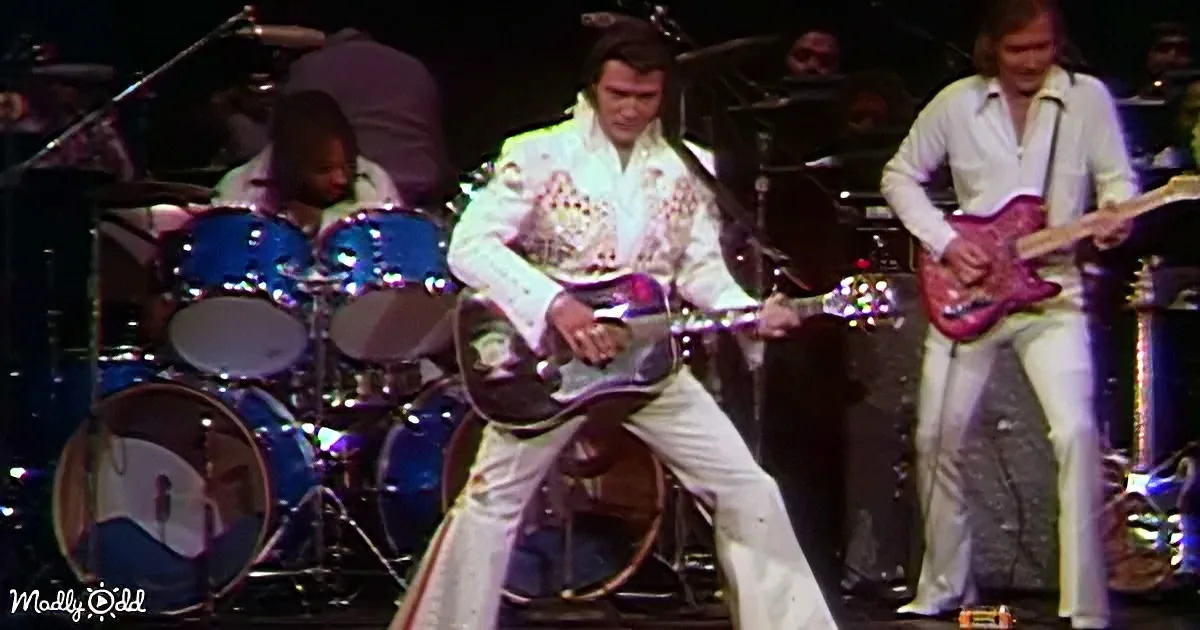Elvis Presley in white jumpsuit plays guitar on stage with band.