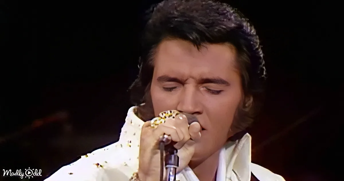 A man in a white sequined outfit sings into a microphone, his eyes closed, against a dark background.