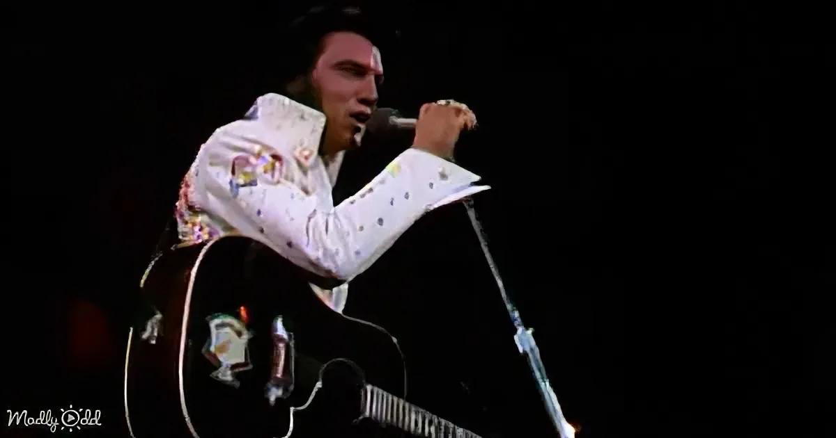 Elvis Presley in white shirt, holding guitar, singing into microphone on stage.