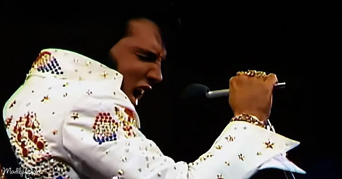 A performer in a white sequined jacket sings into a microphone, holding it with a gold chain.