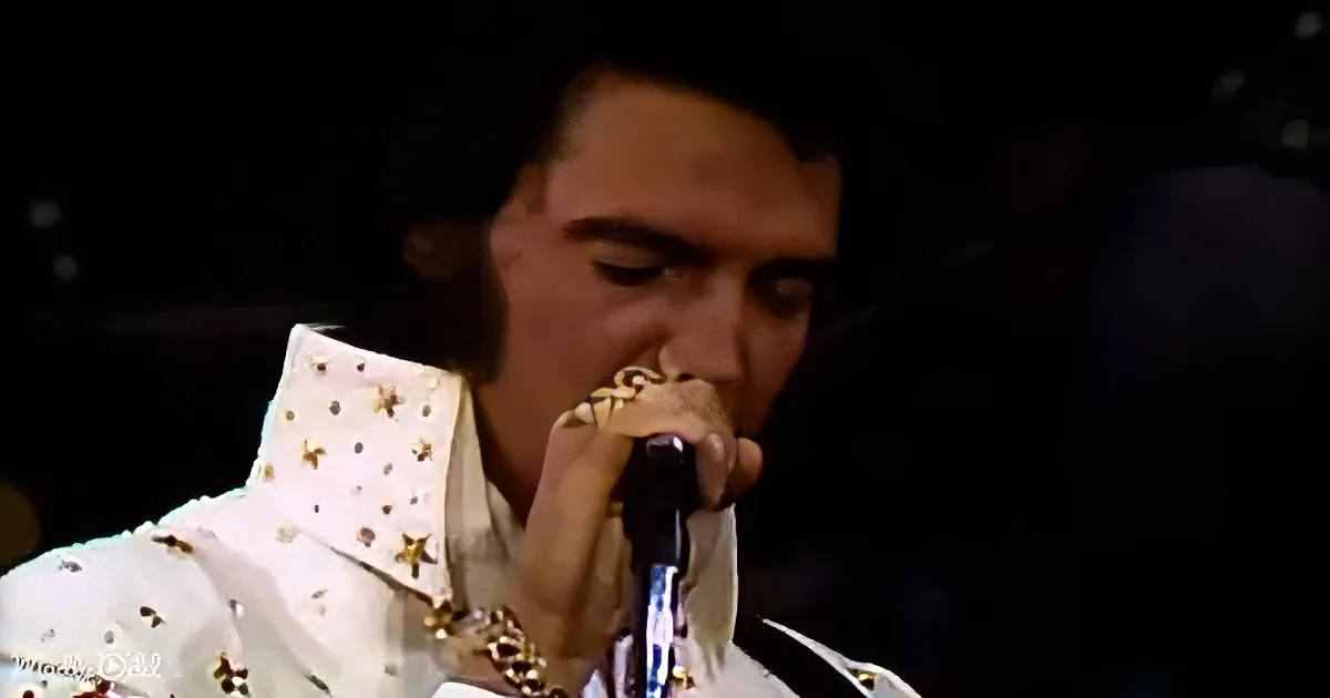 A performer in a white sequined suit sings into a microphone, wearing a gold chain.