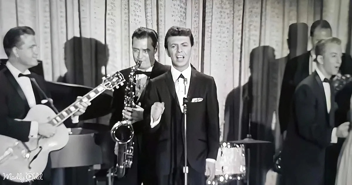 A man in a suit sings into a microphone, surrounded by musicians playing instruments.