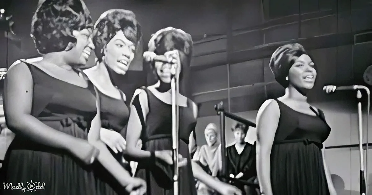 Four women in black dresses sing on stage, smiling and holding microphones.