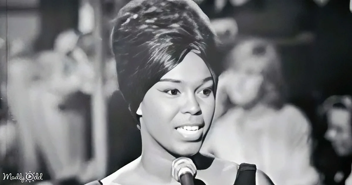 A woman with a short, textured hairstyle sings into a microphone, smiling, with a blurred audience in the background.