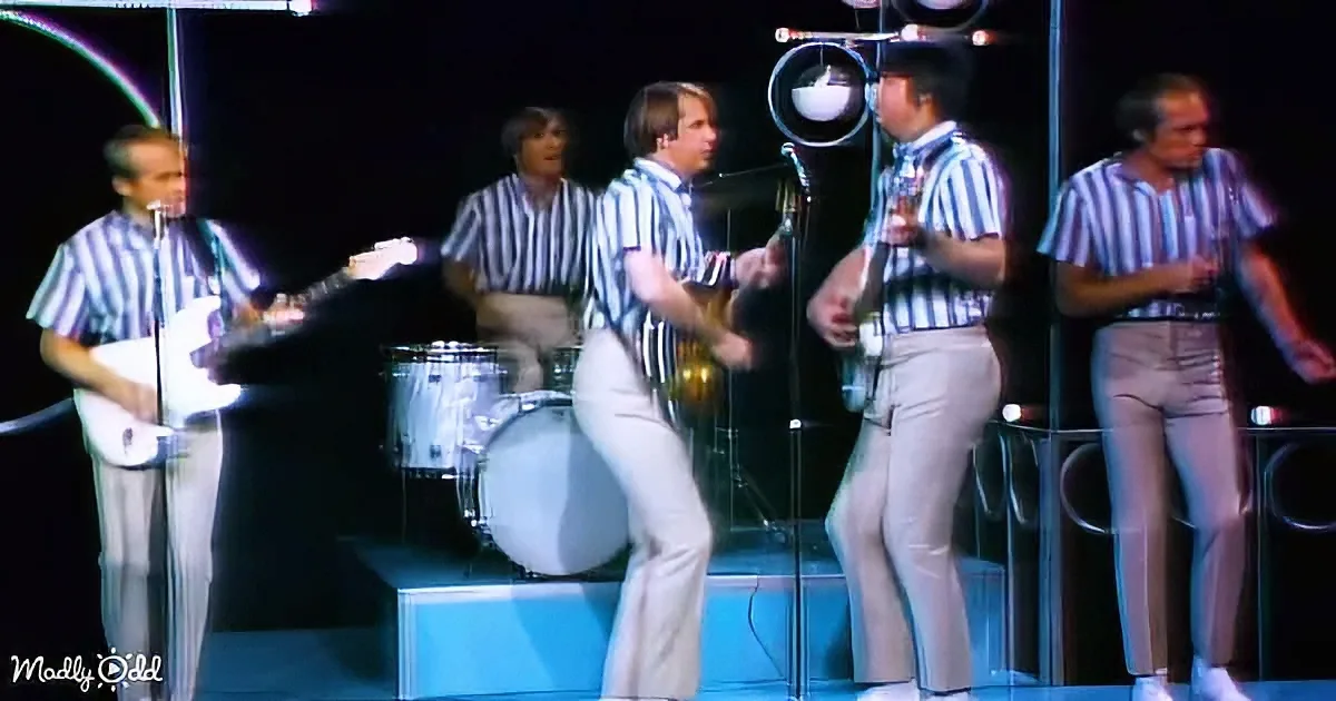 Musicians in striped shirts and white pants perform on stage with guitars and drums.