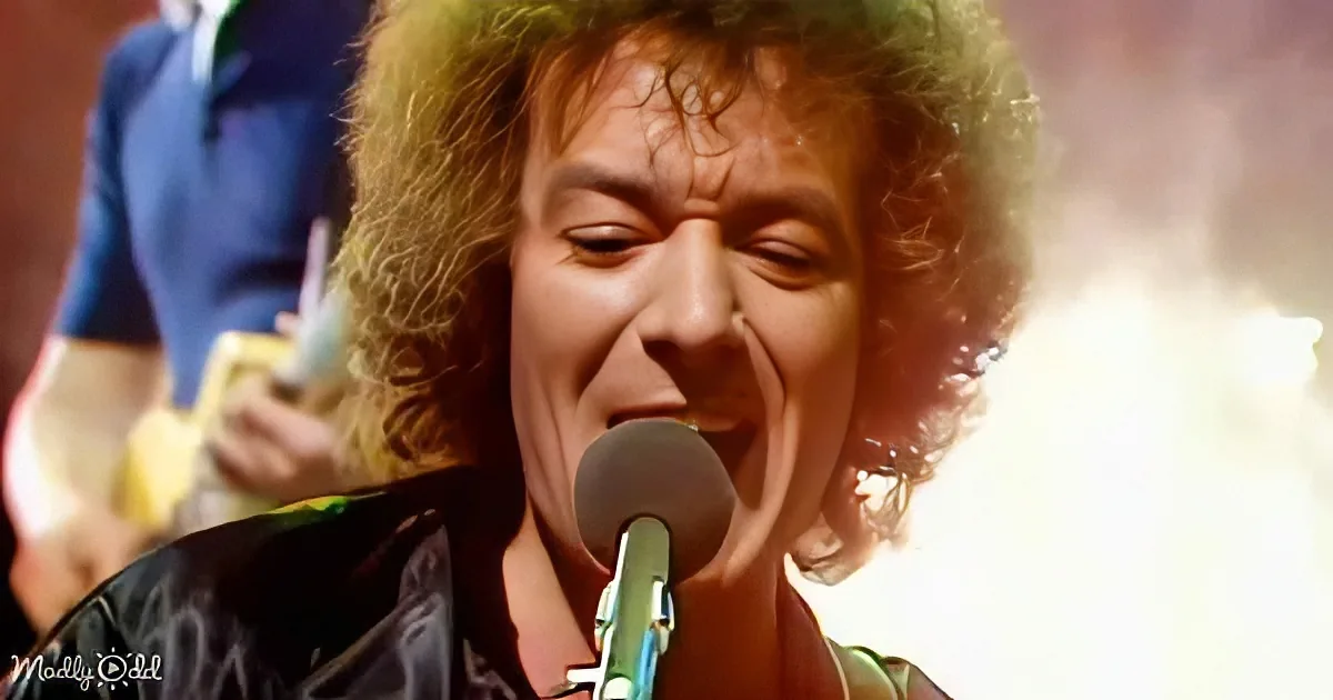 A person with curly hair singing into a microphone, wearing a dark shirt, with a guitarist in the background.