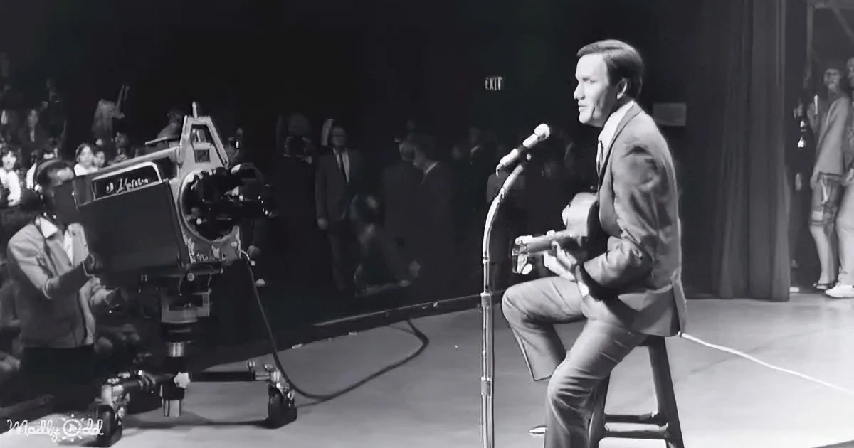 A man in a suit plays guitar on stage, with a camera operator and audience in the background.