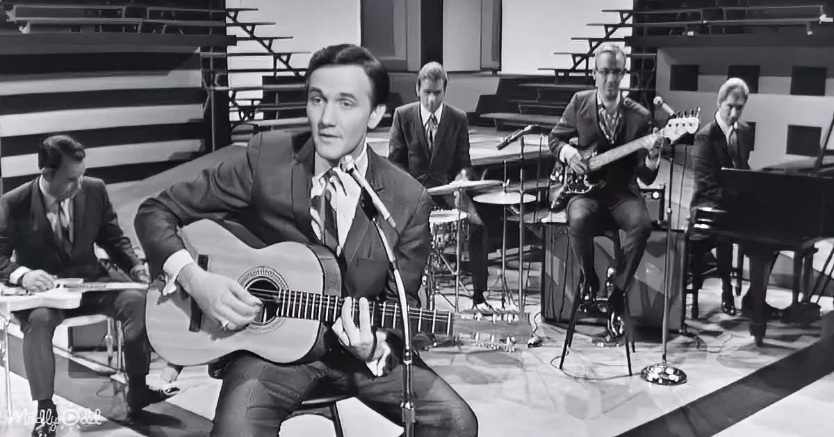 A man in a suit plays guitar on stage, surrounded by musicians and instruments.