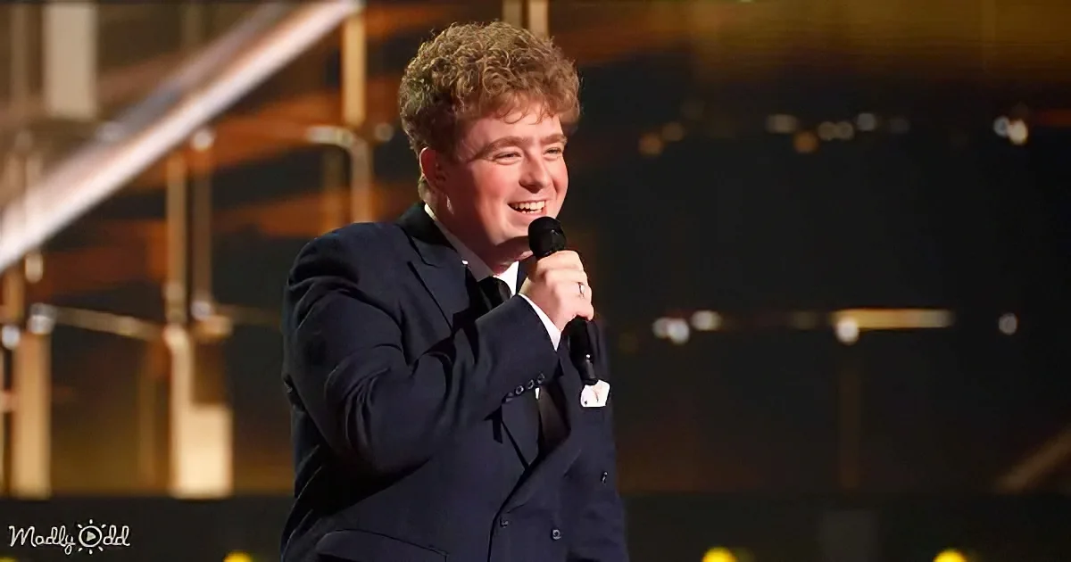 A man in a dark suit smiles while holding a microphone on stage.