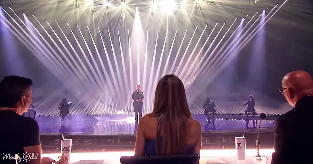 A person with long hair sits in front of a stage with bright lights and performers.