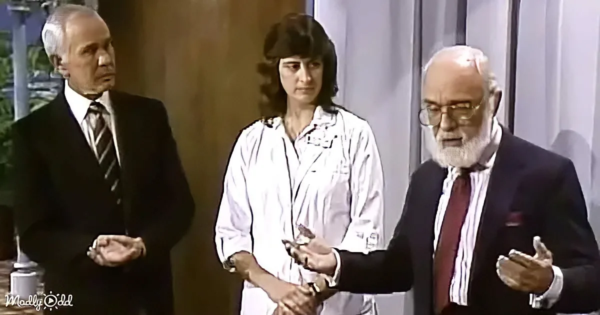 A man in a suit gestures while holding a small object, with another man in a suit and a woman in a lab coat standing nearby.