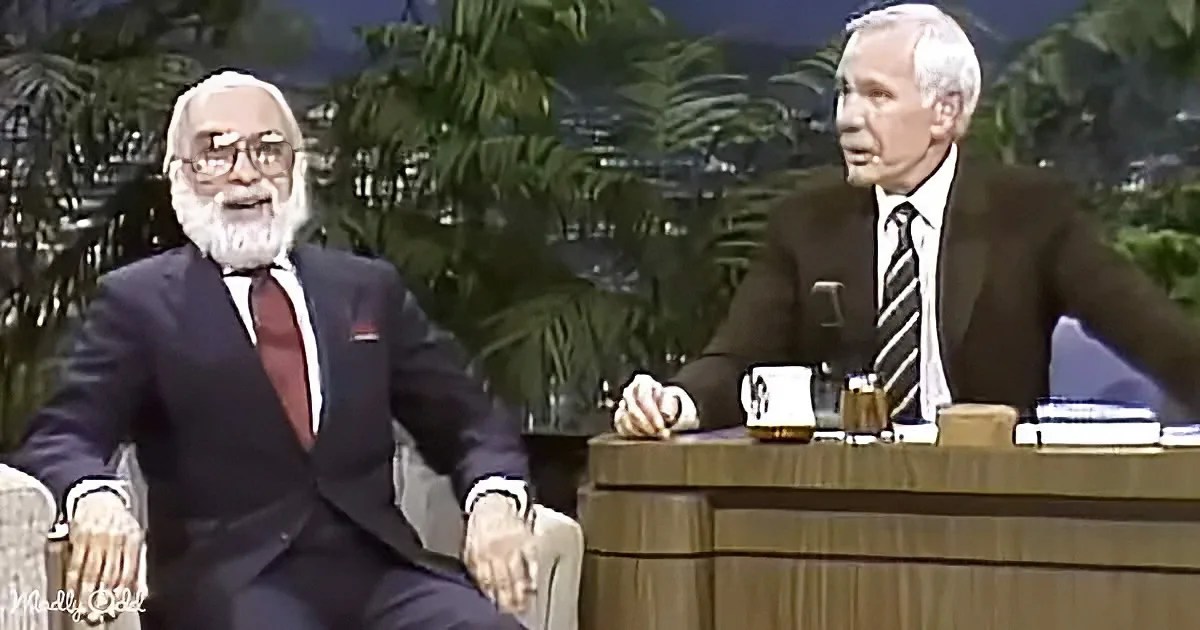 An older man in a suit sits on a couch, smiling, while another man in a suit sits at a desk with a microphone.
