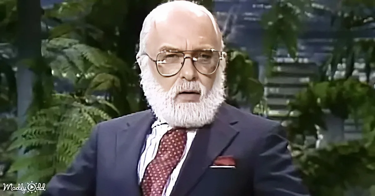 An older man with a white beard and glasses, wearing a suit and tie, stands in front of greenery.
