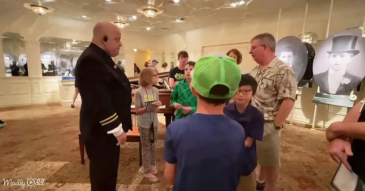 A man in a navy uniform speaks to a group of children and adults in a museum setting.