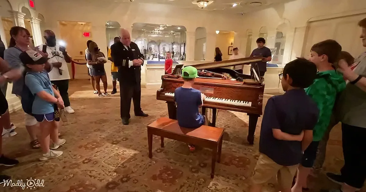 A young boy in a green cap plays a grand piano as a group of children and adults watch in a museum-like setting.