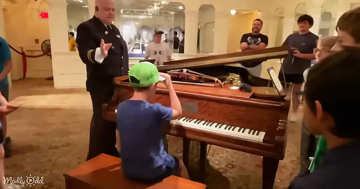 A young boy in a green cap plays a grand piano while a man in a uniform gestures nearby, surrounded by onlookers.