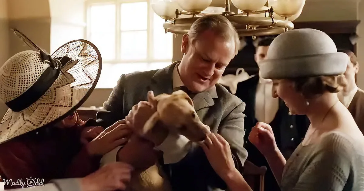 A man holds a small dog surrounded by people in vintage attire, with a chandelier above.