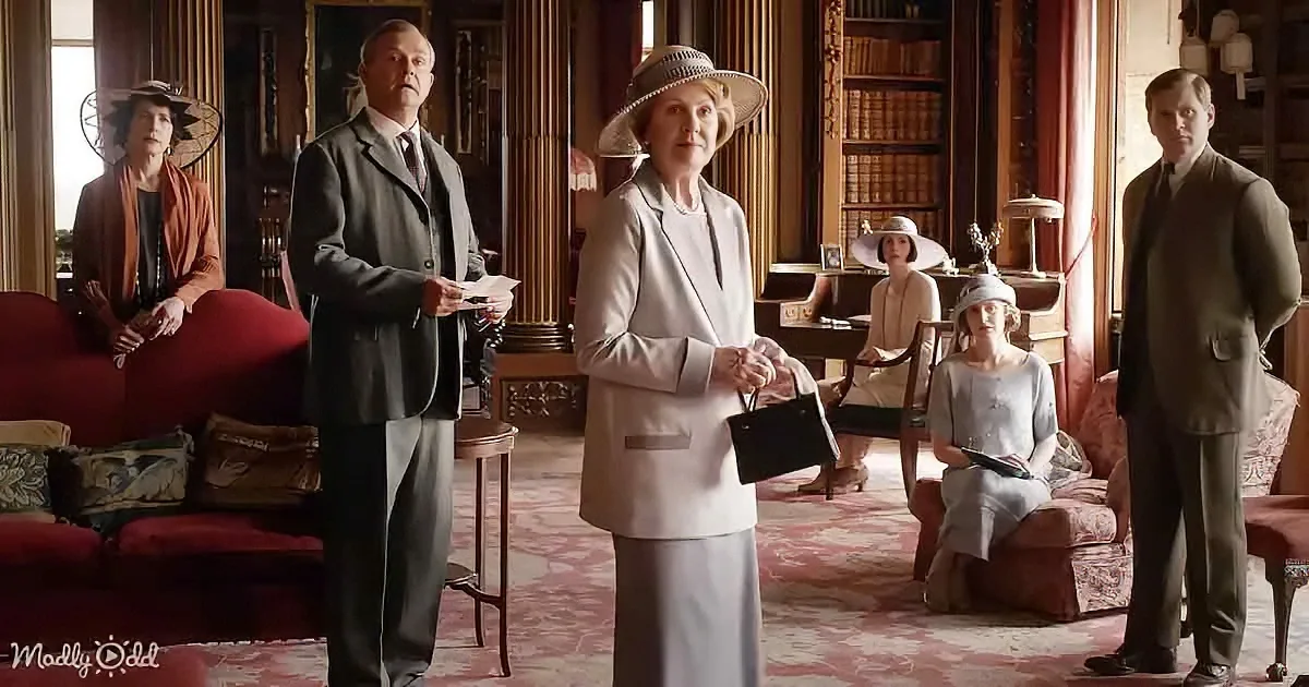 A woman in a light gray suit and hat stands in a grand room, holding a small black purse, surrounded by others in formal...