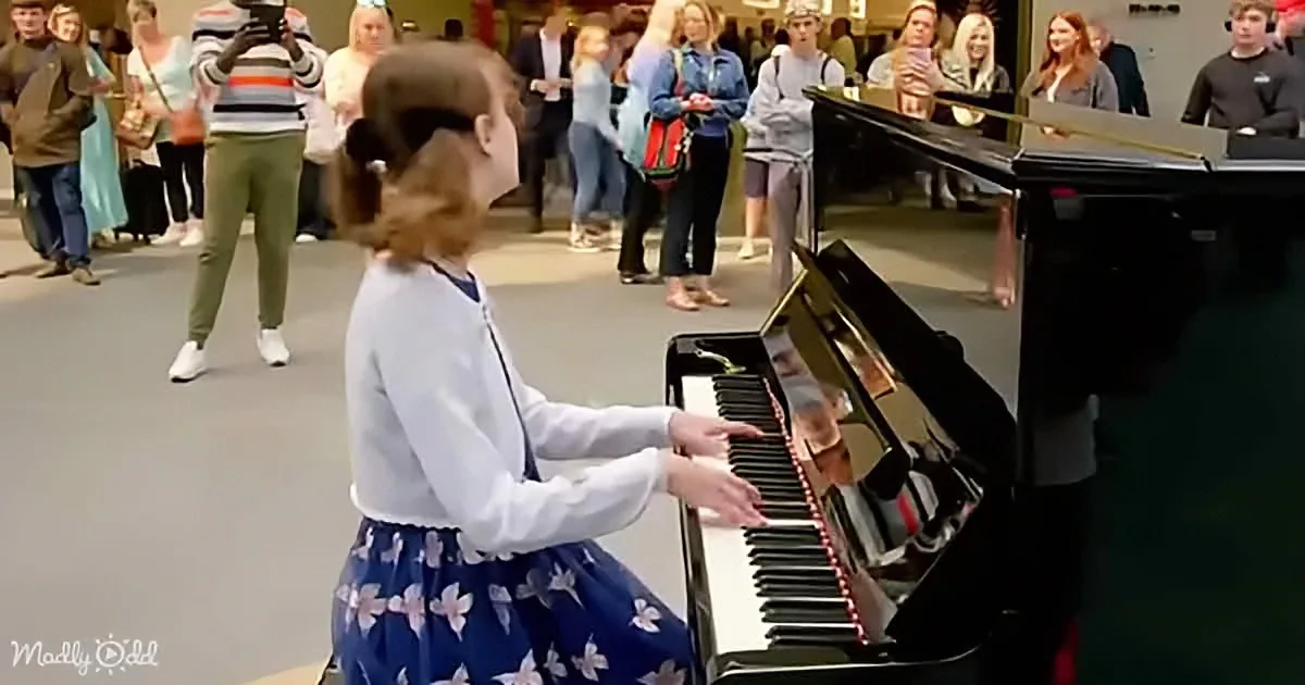 A young girl in a blue floral dress plays a grand piano in front of a crowd of onlookers.