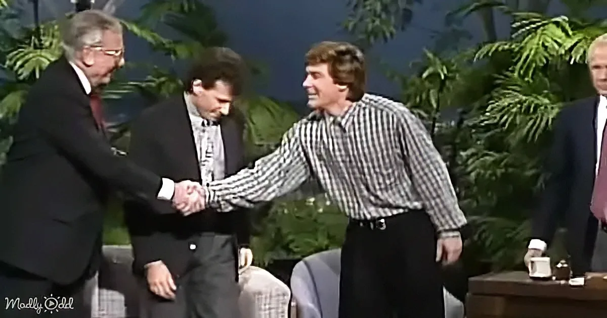A man in a checkered shirt shakes hands with another man in a suit, surrounded by greenery and other people.