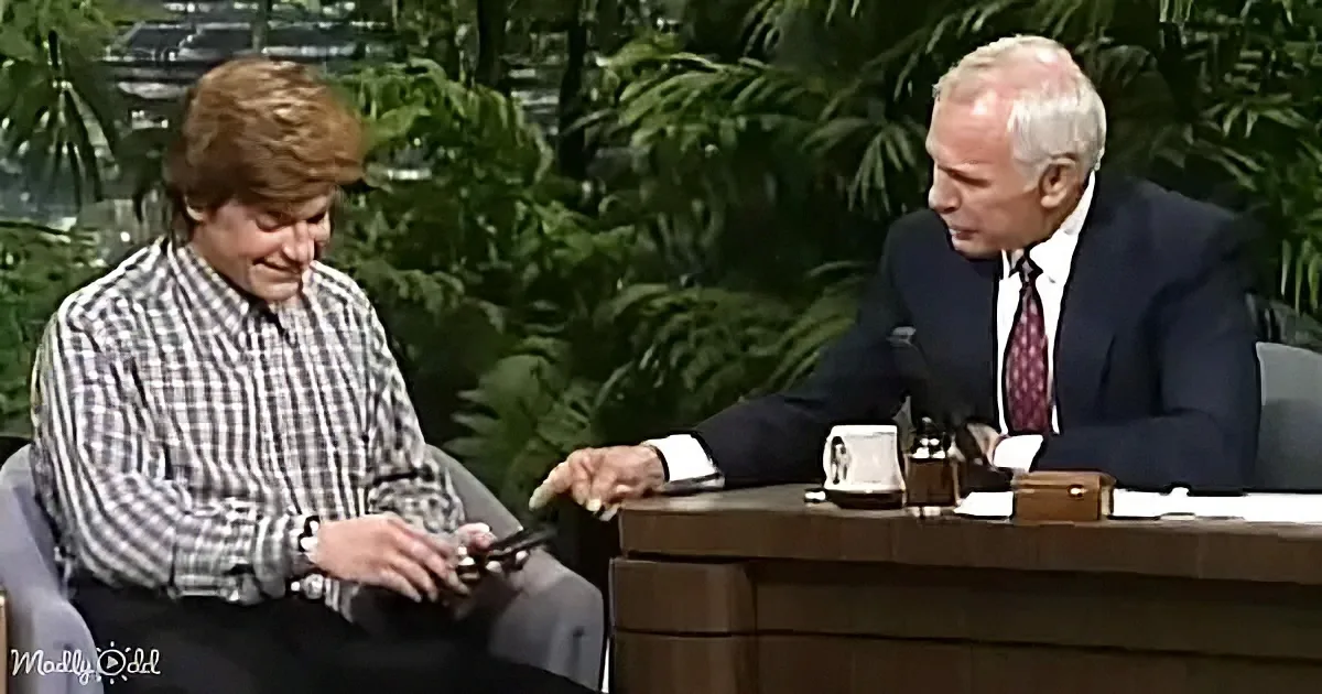 A man in a plaid shirt sits on a couch, holding a phone, while another man in a suit gestures with his hand.
