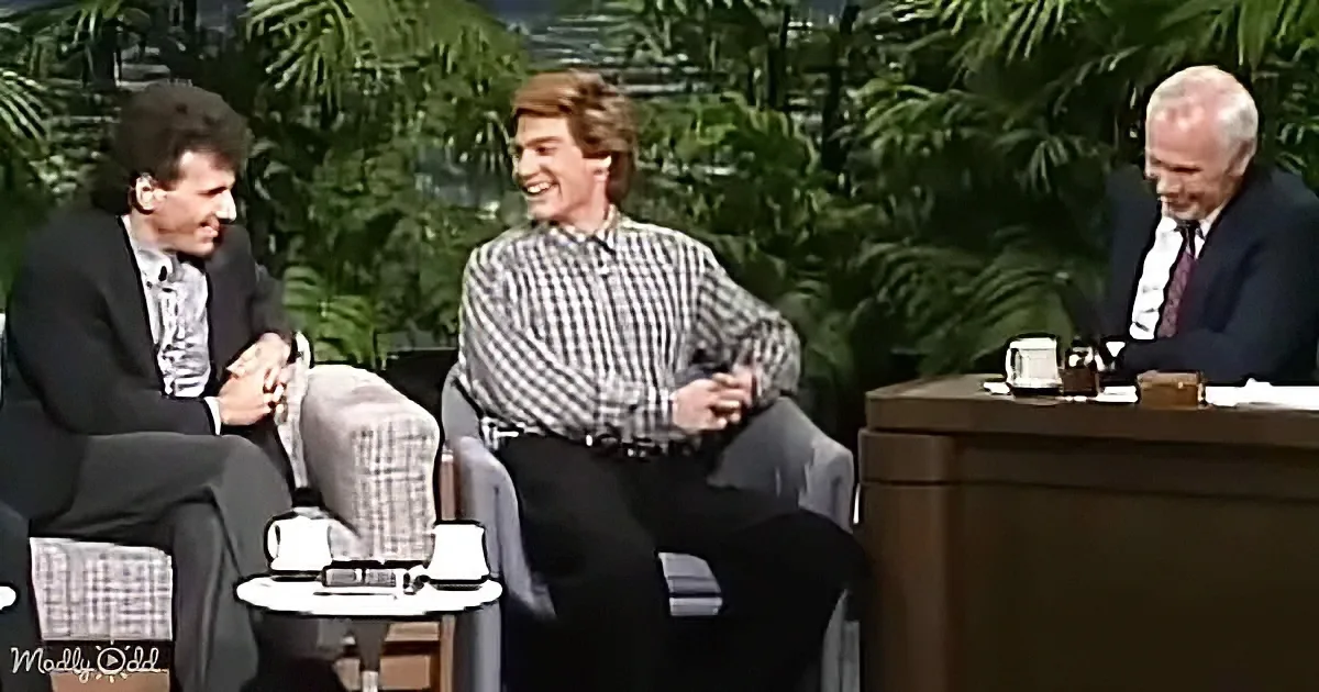 Three men are seated on a talk show set, with one in a plaid shirt laughing and another in a suit, both appearing engaged...