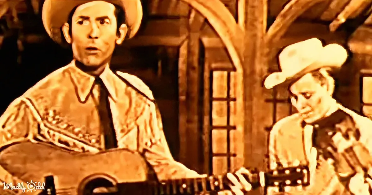 A man in a cowboy hat plays guitar, another man in a white hat looks on in a rustic setting.