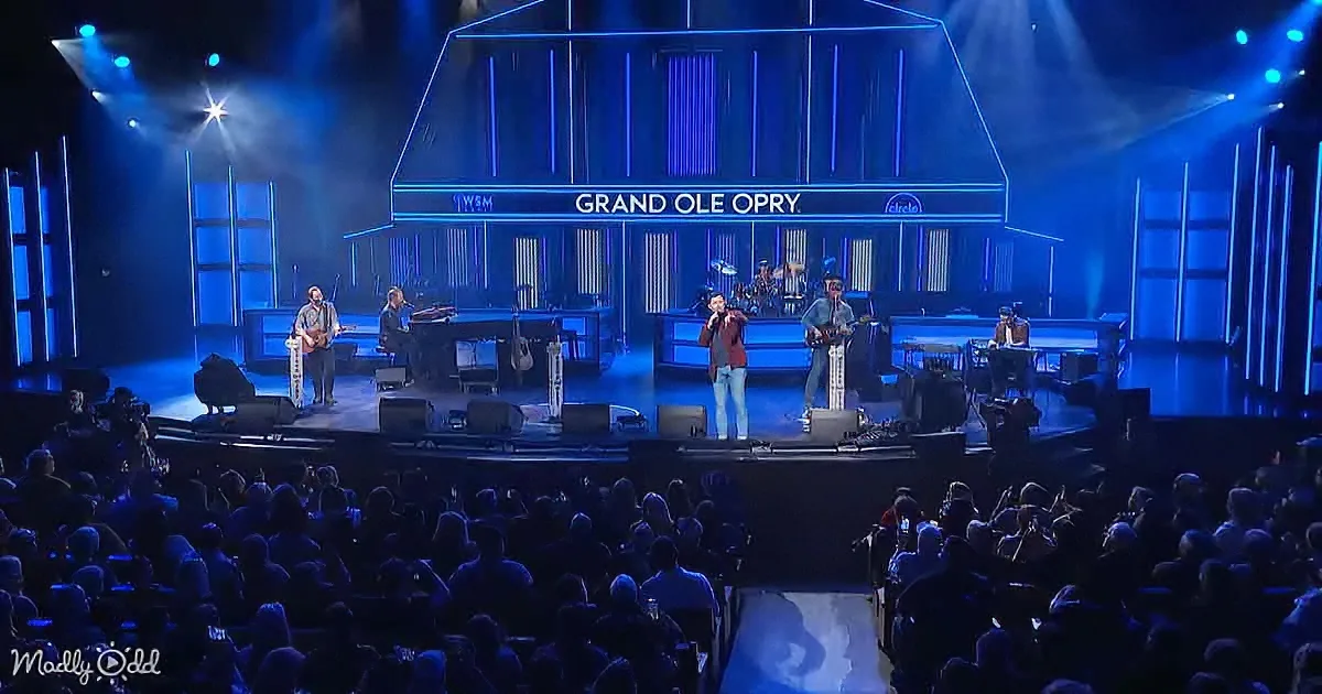 A band performs on stage at the Grand Ole Opry with blue lighting and a large audience.