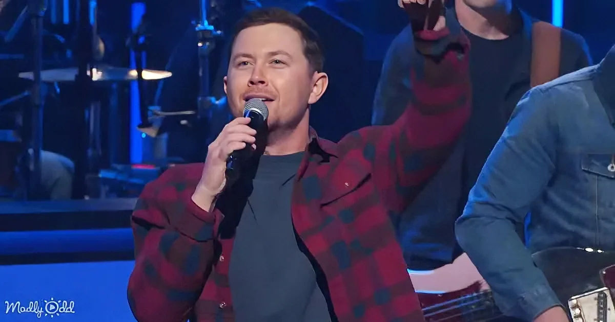 A man in a red plaid jacket sings into a microphone on stage with a band.