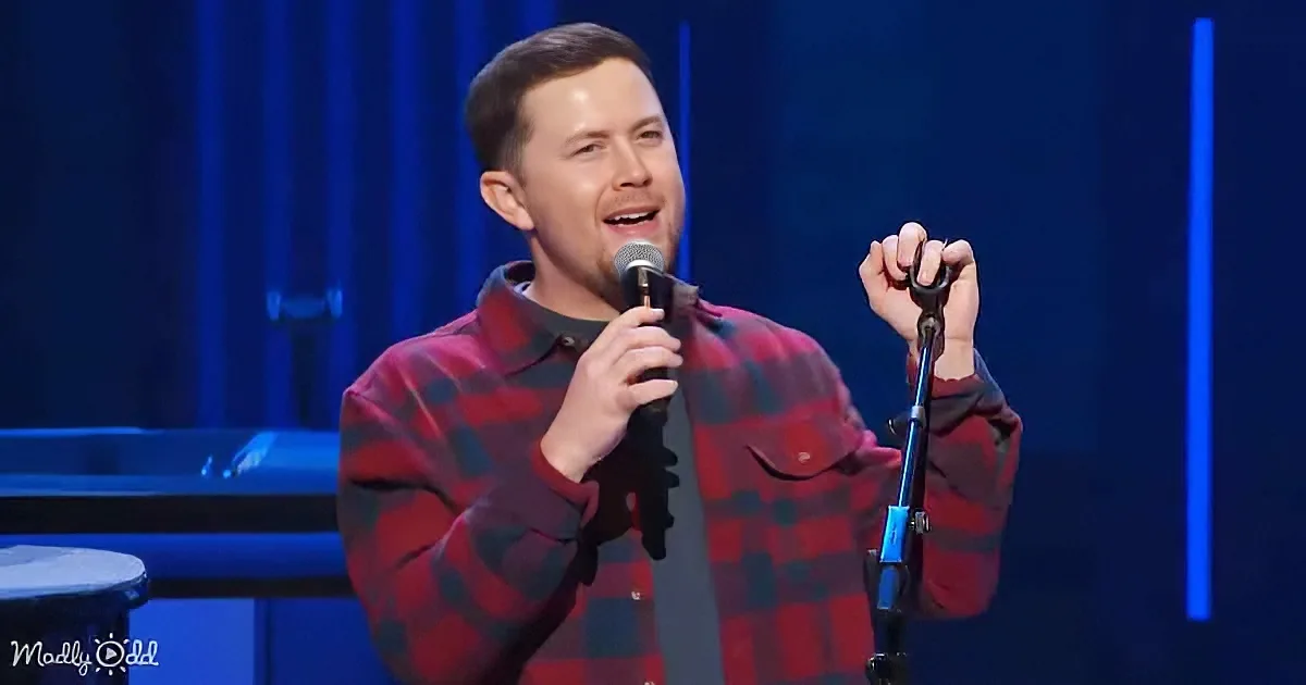 A man in a red plaid shirt holds a microphone, singing on stage with a blue-lit background.