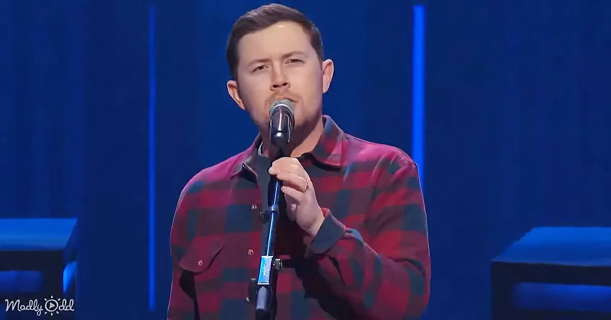A man in a red plaid shirt sings into a microphone on stage with blue lighting.