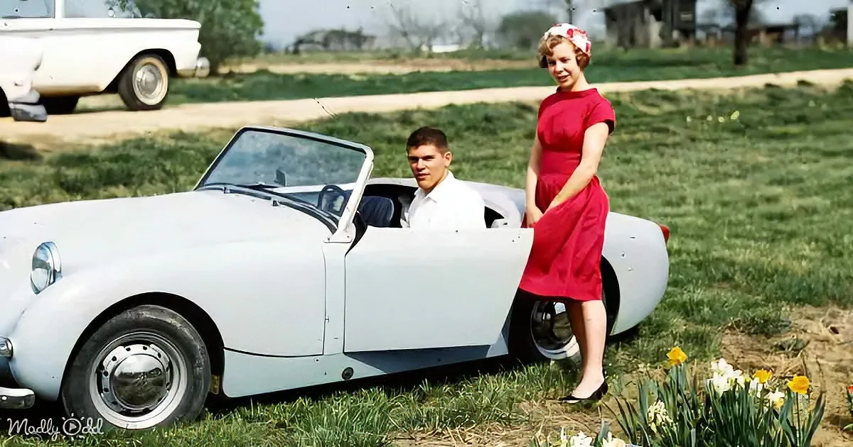 A man and woman pose beside a white vintage convertible car in a grassy field with daffodils.