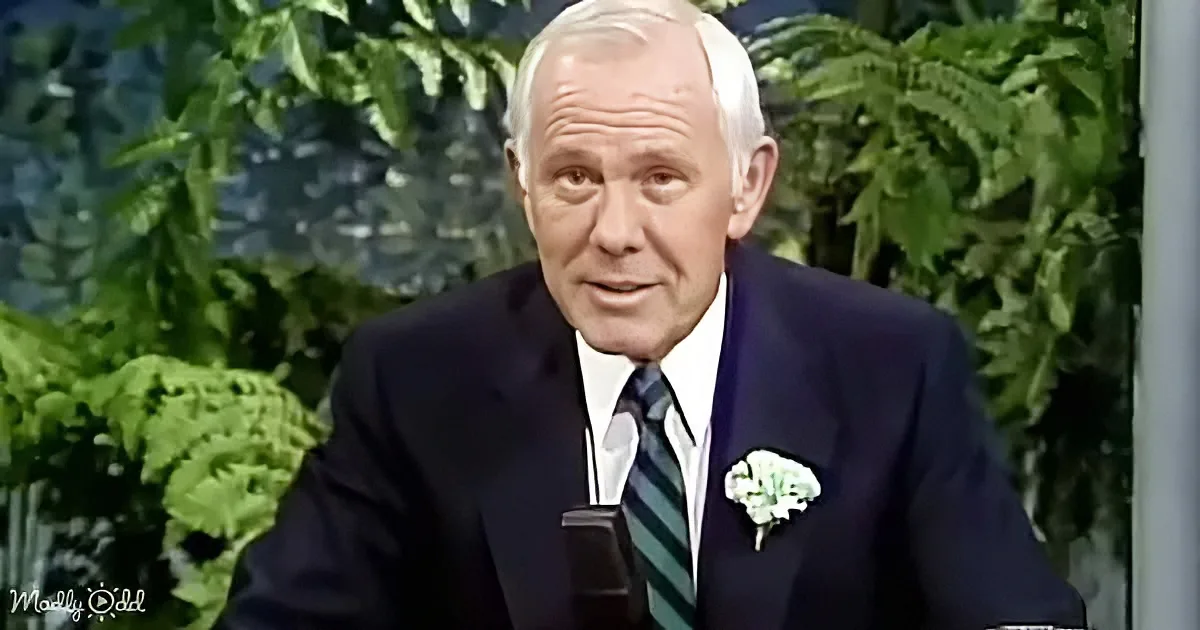 An older man in a suit and tie speaks into a microphone, with green foliage in the background.