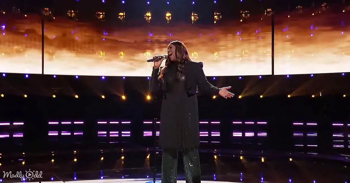 A person in black attire sings passionately on stage with colorful lights and a reflective floor.