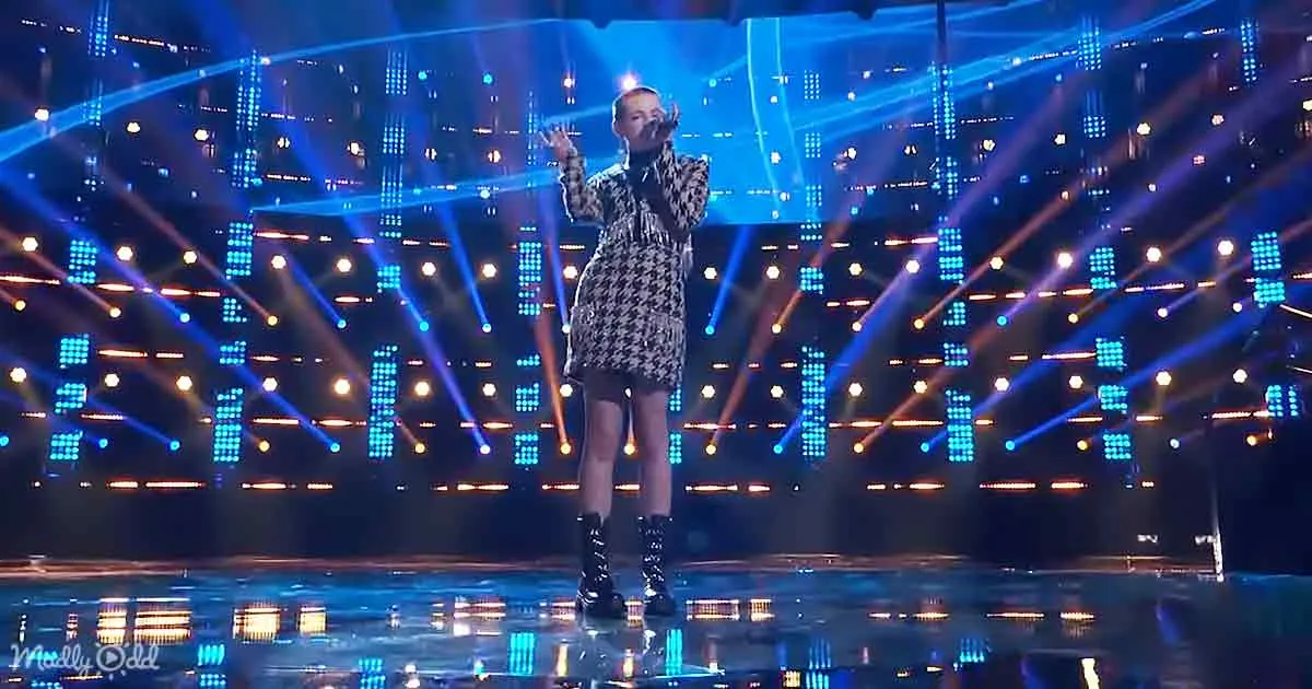 A person in a houndstooth dress and boots stands on a stage with blue and orange lights.