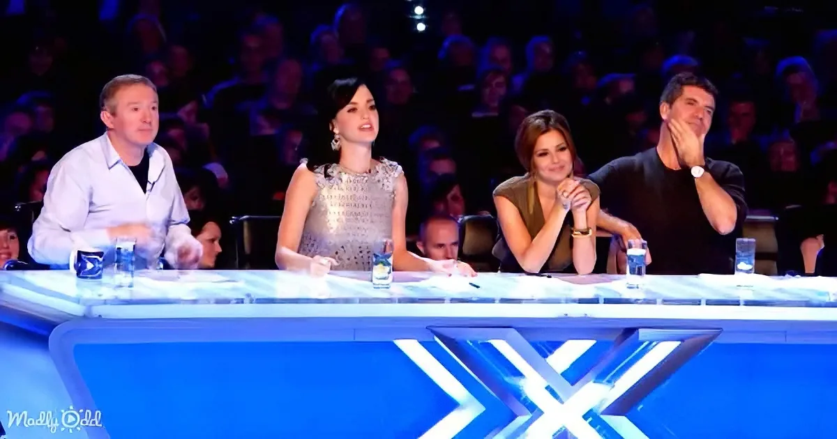 Four judges sit at a table with glowing blue lights, clapping and watching an audience.