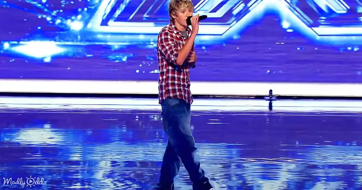 A young man in a plaid shirt sings on stage with a microphone, illuminated by blue lights.