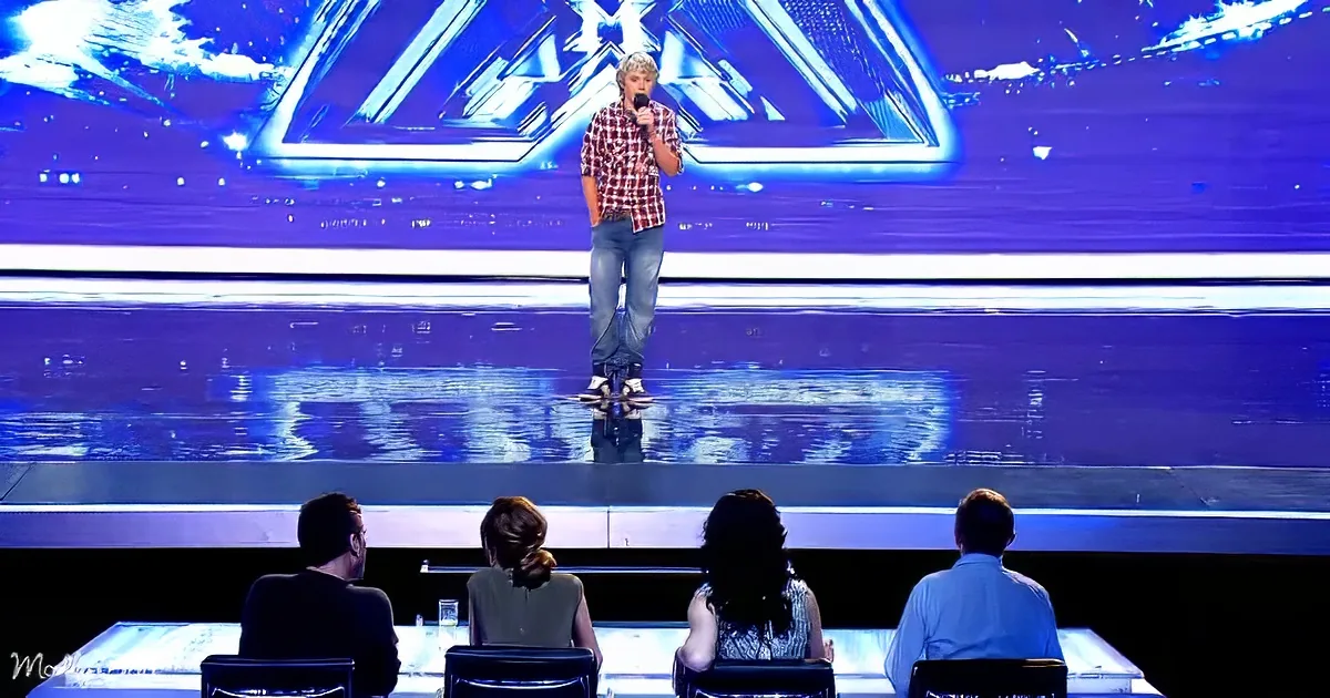 A young man in a plaid shirt stands on a stage with a microphone, addressing judges seated at a table.