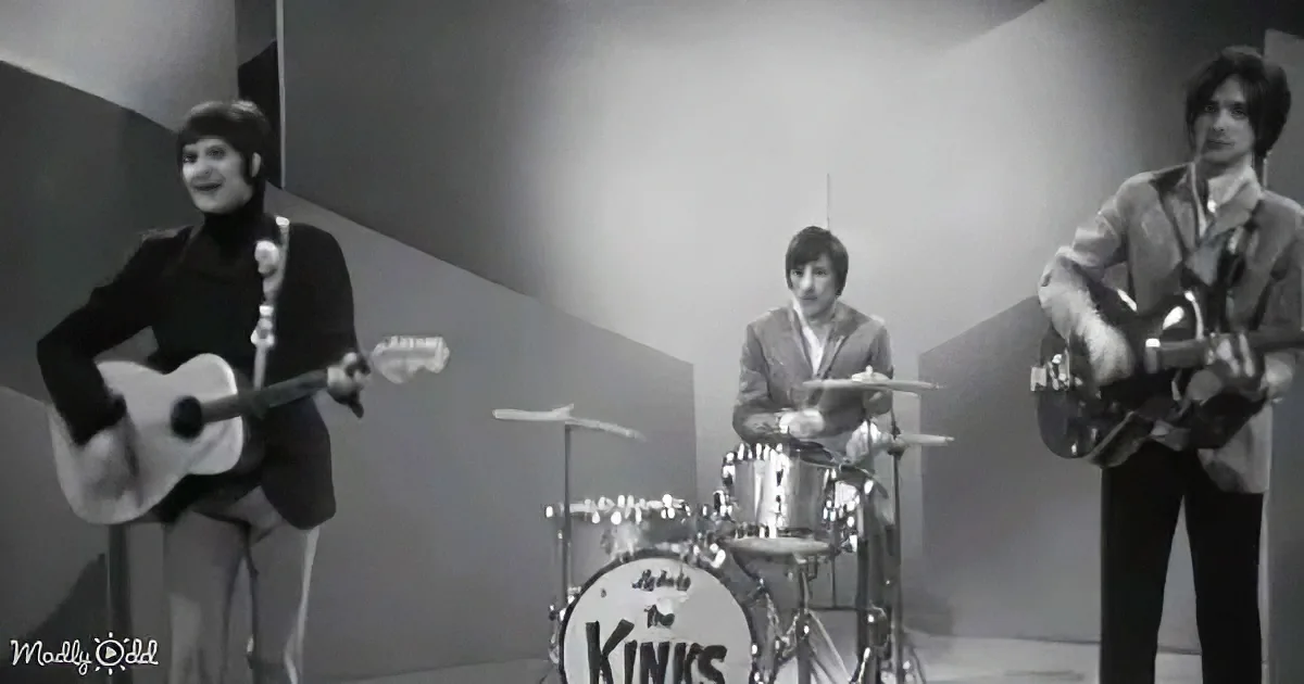 The Kinks band performs with a drummer, guitarist, and bassist in a vintage black-and-white setting.