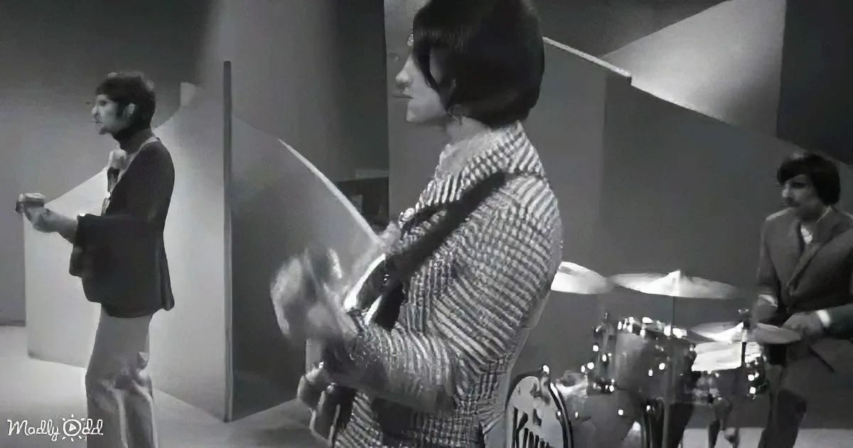 A woman plays guitar in a black-and-white scene, with a man singing and another playing drums in the background.