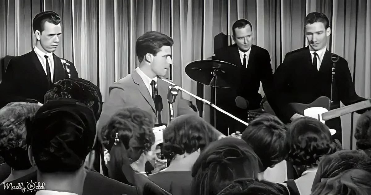 A man in a suit sings into a microphone, surrounded by a band and an audience.