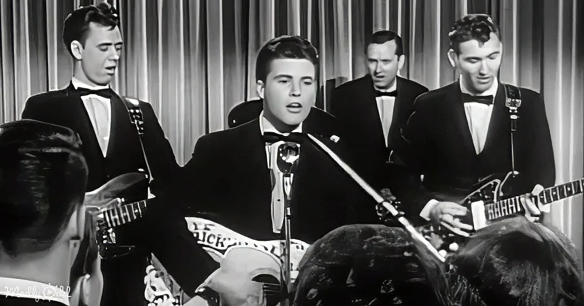 A man in a tuxedo sings into a microphone, accompanied by three musicians in suits playing guitars.