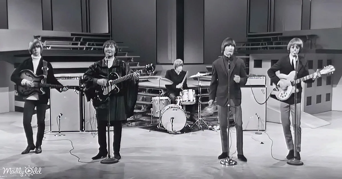 A band performs on stage with guitars, drums, and microphones, dressed in suits, in a black-and-white setting.
