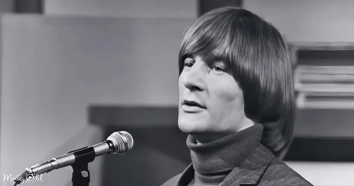 A person with shoulder-length hair sings into a microphone, wearing a turtleneck sweater.