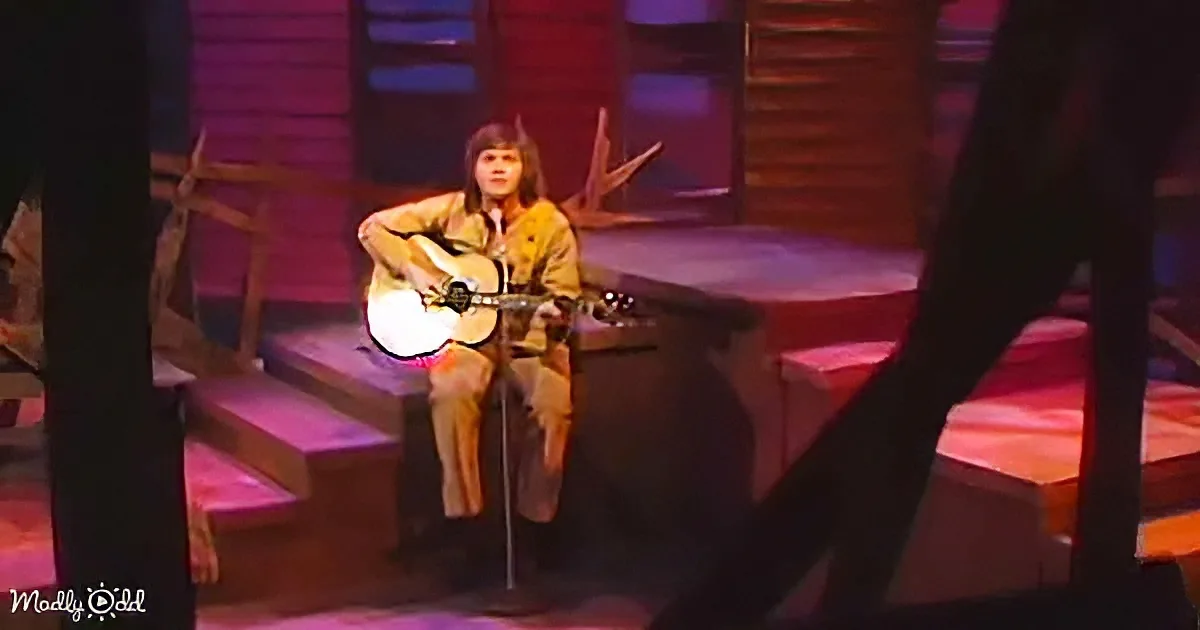 A person sits on a stage, playing an acoustic guitar, with a microphone in front of them.