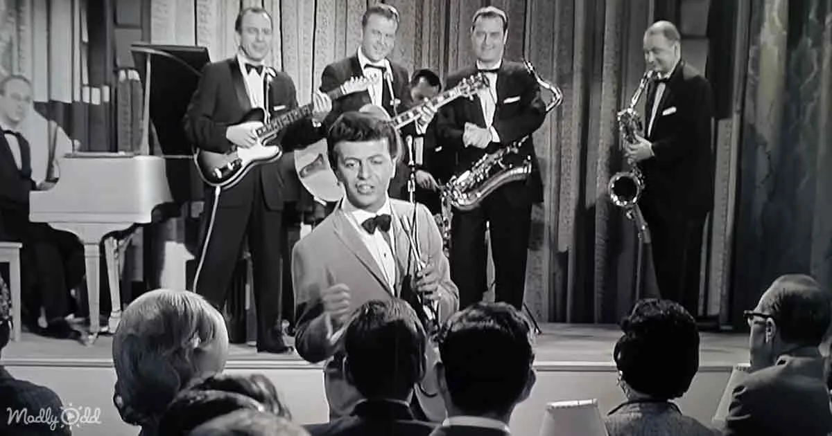 A man in a suit sings on stage with a band, surrounded by musicians and an audience.