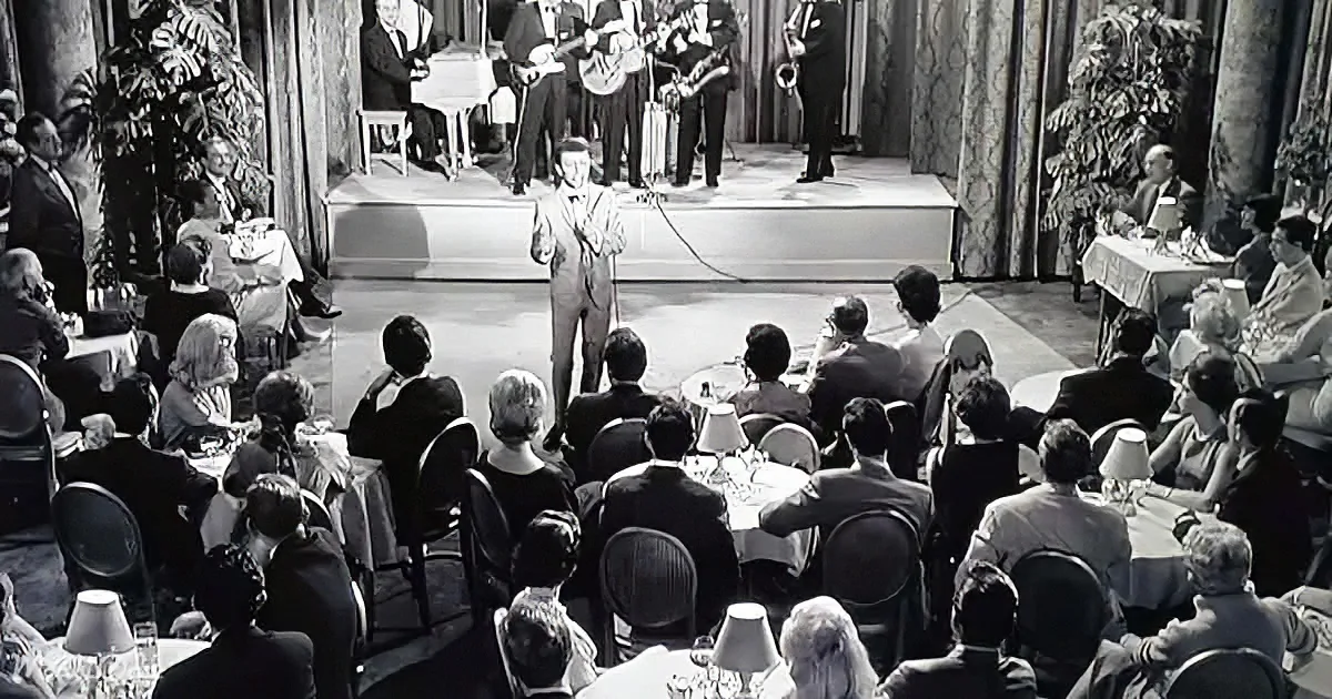 A man in a suit stands on stage, addressing an audience seated at tables with lamps.