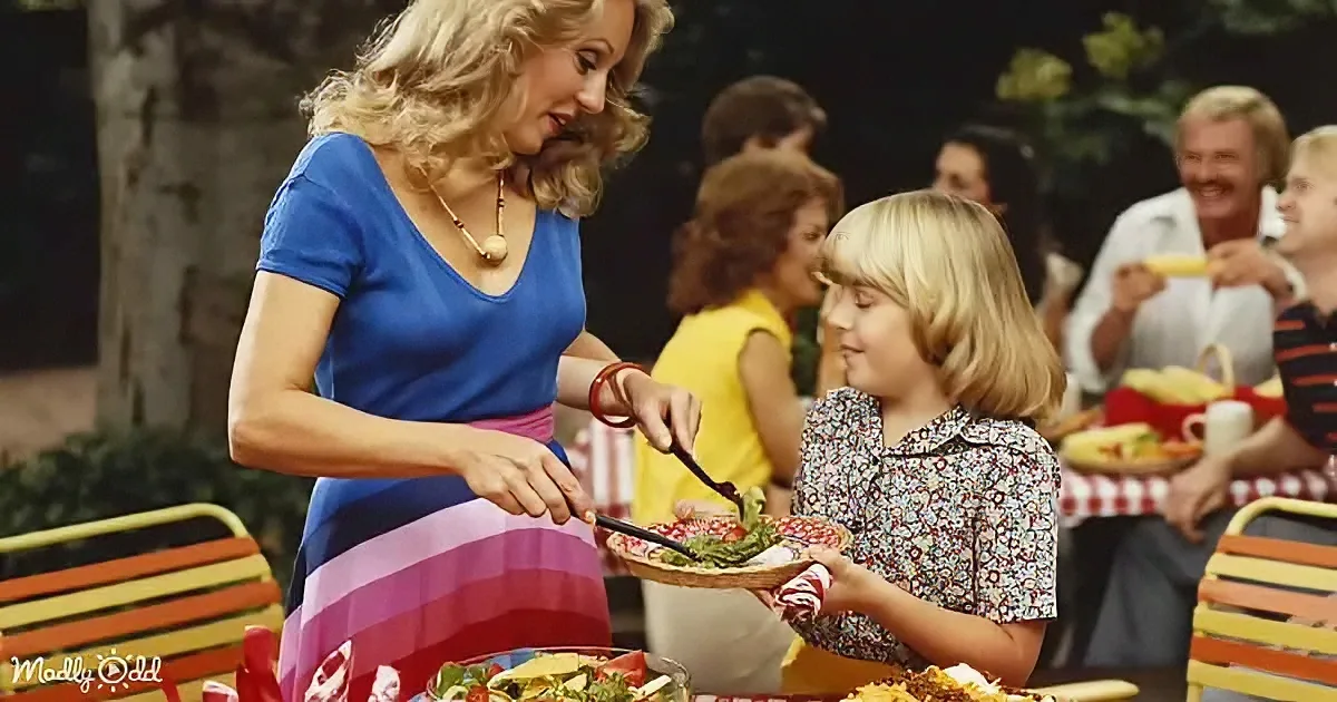 A woman in a blue dress serves salad to a young girl at a picnic, surrounded by other people enjoying food.