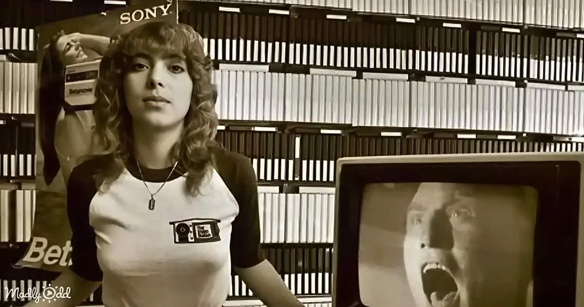 A woman in a white t-shirt with a camera graphic stands in front of a Sony Betamax advertisement and a TV showing a person.